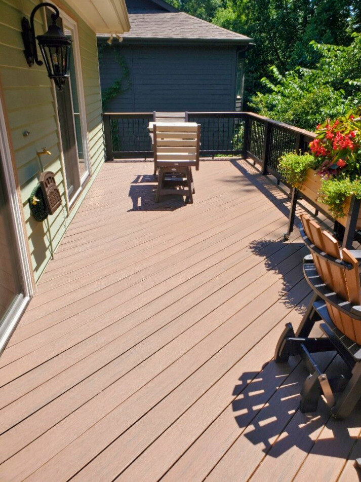 A close-up view of a spacious brown composite deck with diagonal planks. Two wooden chairs are placed on the deck, and a planter box with red flowers sits near the railing on the right.